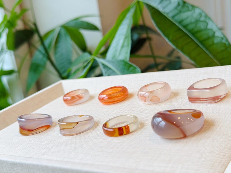 May include: A collection of eight smooth, polished rings crafted from translucent banded agate stone. Each ring features unique natural color variations, including shades of orange, white, brown, and grey, with distinct swirling patterns and polished surfaces.