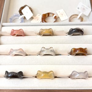 Puede incluir: Una exhibición de varios anillos de piedras preciosas en una caja de joyería de madera. Los anillos presentan una variedad de colores y diseños, incluyendo formas de lazo y formas irregulares. Algunos anillos tienen etiquetas de precio. Los anillos se exhiben sobre una superficie blanca.