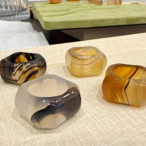 May include: Four polished stone rings in shades of brown, grey, and cream. Each ring has a unique pattern and shape, with a smooth, rounded design. The rings are displayed on a neutral-coloured surface.