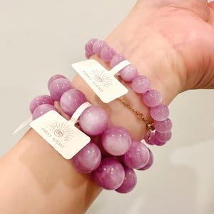 May include: Two bracelets with large, round, light purple beads are worn on a wrist. Each bracelet has a tag that reads "FIRST SIGHT" with an eye symbol. One bracelet is connected with a gold chain.