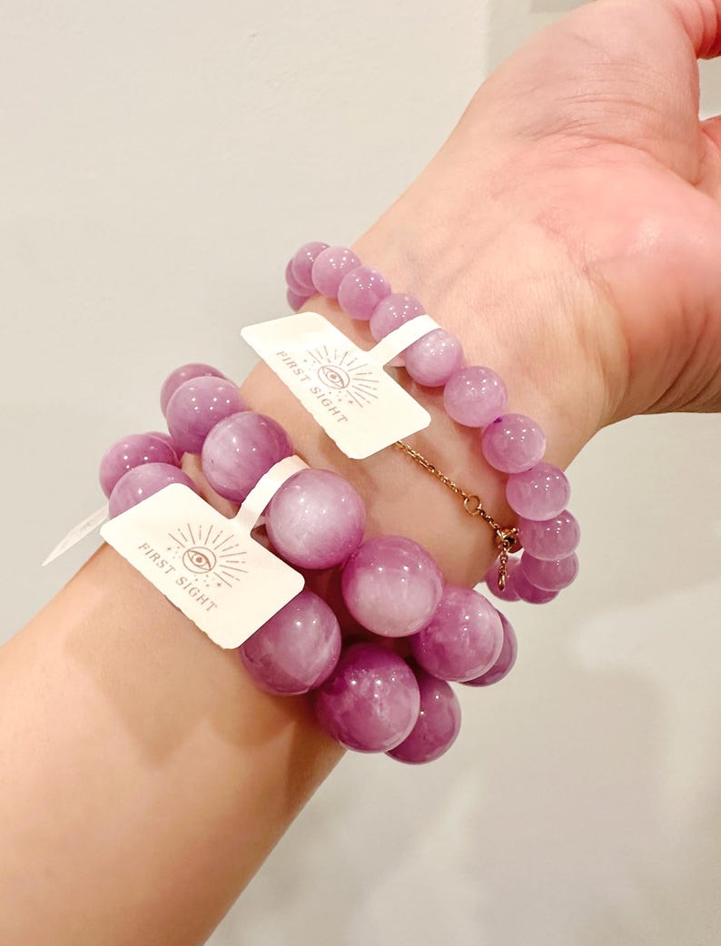 May include: Two bracelets with large, round, light purple beads are worn on a wrist. Each bracelet has a tag that reads "FIRST SIGHT" with an eye symbol. One bracelet is connected with a gold chain.
