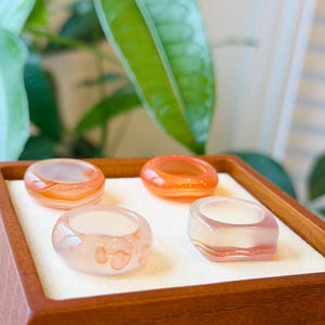 May include: Four rings crafted from translucent stone, each featuring distinct shapes and color variations. Two rings are circular with orange and white banding, one is circular with soft pink inclusions, and one is square with pink and white layers.