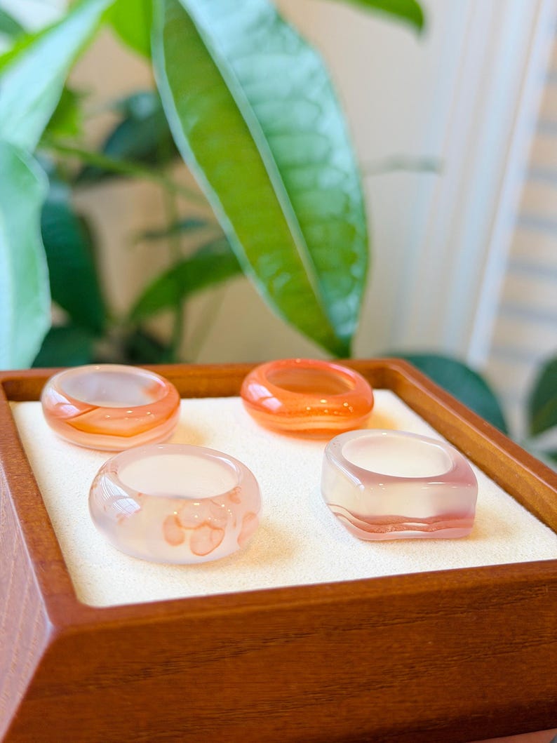 May include: Four rings crafted from translucent stone, each featuring distinct shapes and color variations. Two rings are circular with orange and white banding, one is circular with soft pink inclusions, and one is square with pink and white layers.