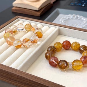 Op de afbeelding: Twee armbanden met barnsteen en heldere kwarts kralen. De armbanden worden gepresenteerd in een houten bakje met een crèmekleurig interieur. De kralen hebben een mix van oranje, gele en heldere tinten.
