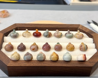 May include: A wooden tray displays a collection of small, carved stone cat figurines. The cats are in various colors, including brown, tan, and green. Each figurine has a rounded body and a small, raised head. The tray is lined with a cream-colored fabric.