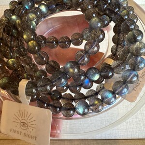 May include: A close-up of a dark grey labradorite gemstone beaded necklace with iridescent blue and green hues. The necklace is wrapped around a clear glass bowl. The necklace is labelled "First Sight".
