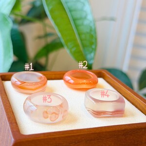 May include: Four smooth, translucent resin rings arranged in a square formation. Ring #1 is peach-colored, #2 is bright orange, #3 is pale pink with darker pink spots, and #4 is a square-shaped ring with a pale pink and white gradient.
