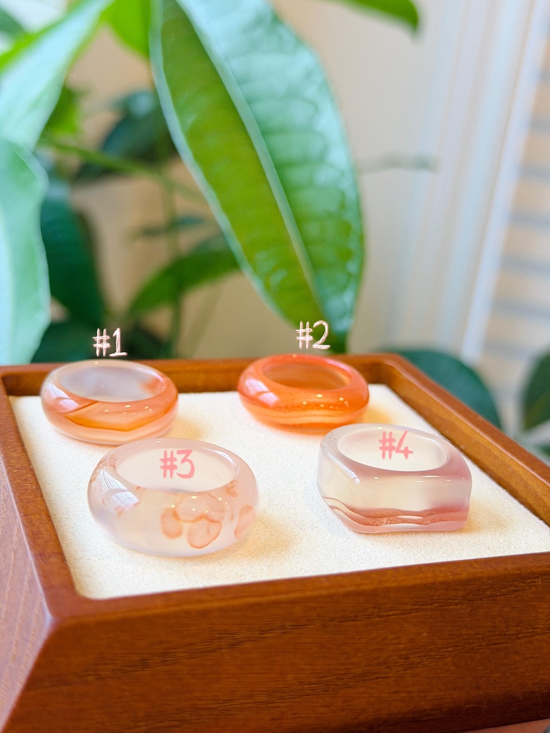 May include: Four smooth, translucent resin rings arranged in a square formation. Ring #1 is peach-colored, #2 is bright orange, #3 is pale pink with darker pink spots, and #4 is a square-shaped ring with a pale pink and white gradient.