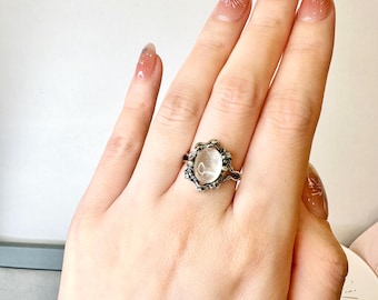 May include: A silver ring with a clear oval-shaped gemstone set in a detailed, ornate design. The ring is on a person's finger.