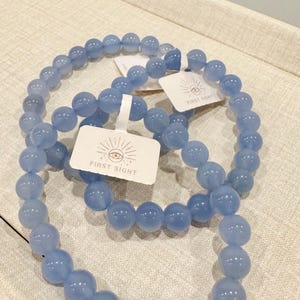 May include: Three light blue beaded bracelets. Each bracelet is made of round, polished beads. Each bracelet has a white tag that reads "FIRST SIGHT" with an eye graphic. The bracelets are displayed on a light beige surface.