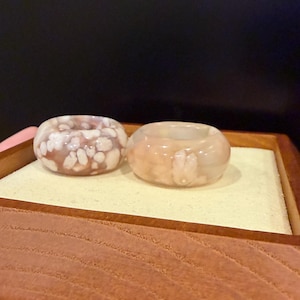 May include: Two gemstone rings in a wooden box. The rings are light pink with white and pink marbling. The rings are round and polished. The box has a light yellow interior. The rings are likely made of jade or agate.