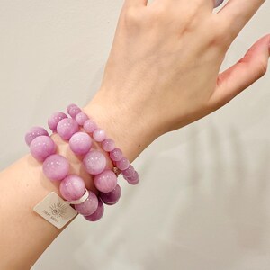 May include: Three beaded bracelets and a ring in varying sizes of light purple. The bracelets are made of large, round beads. A matching ring is worn on a finger. A tag with the text "FIRST SIGHT" is attached to one bracelet.