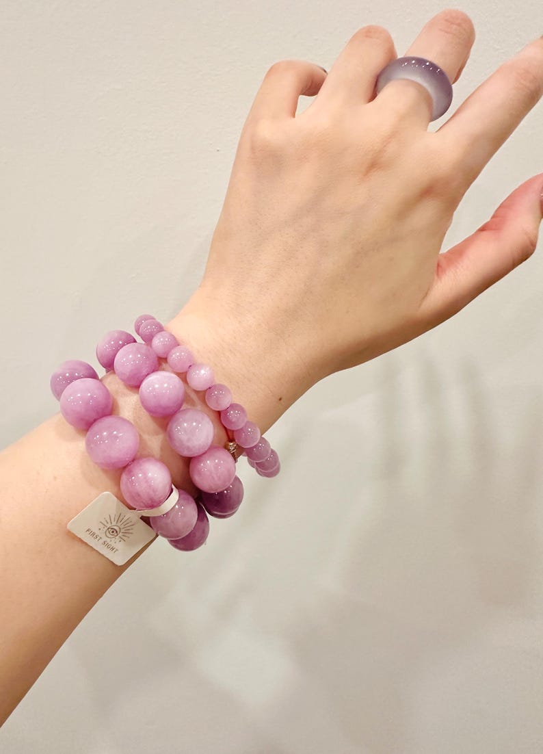 May include: Three beaded bracelets and a ring in varying sizes of light purple. The bracelets are made of large, round beads. A matching ring is worn on a finger. A tag with the text "FIRST SIGHT" is attached to one bracelet.