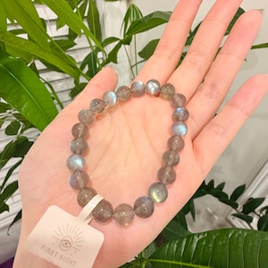 May include: A beaded bracelet made of round, gray labradorite stones. The bracelet is held in a hand, with a white tag attached that reads "FIRST SIGHT" with an eye symbol. The stones have iridescent blue and green flashes.