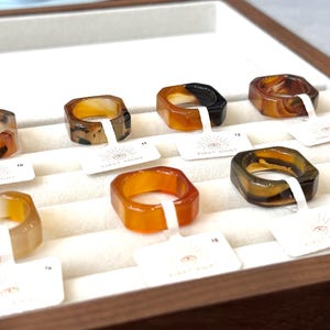 May include: A display of square-shaped rings in various colors, including amber, brown, and black. Each ring is on a white tag with the text "FIRST SIGHT" and a sunburst logo. The rings are arranged in a wooden tray.