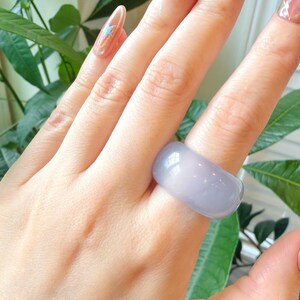 May include: A wide, light purple ring. The ring is smooth and has a rounded shape. The ring is worn on a finger. The background includes green leaves and a glimpse of a fingernail with a star design.