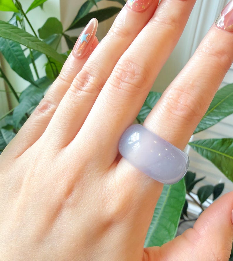 May include: A wide, light purple ring. The ring is smooth and has a rounded shape. The ring is worn on a finger. The background includes green leaves and a glimpse of a fingernail with a star design.