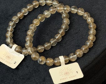 May include: Two stretch bracelets made of round, translucent, brownish-grey rutilated quartz beads with visible golden needle-like inclusions. Each bracelet features a small white rectangular tag printed with an eye icon and the text FIRST SIGHT.