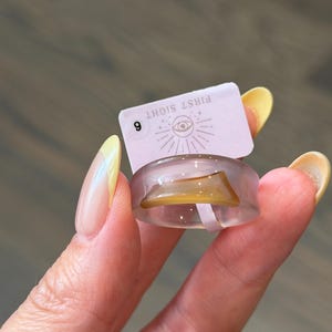 May include: A translucent light purple ring featuring an embedded amber-colored curved accent. Attached to the ring is a small rectangular tag with the text FIRST SIGHT, an eye icon, and the number 6 printed in black ink on a light pink background.
