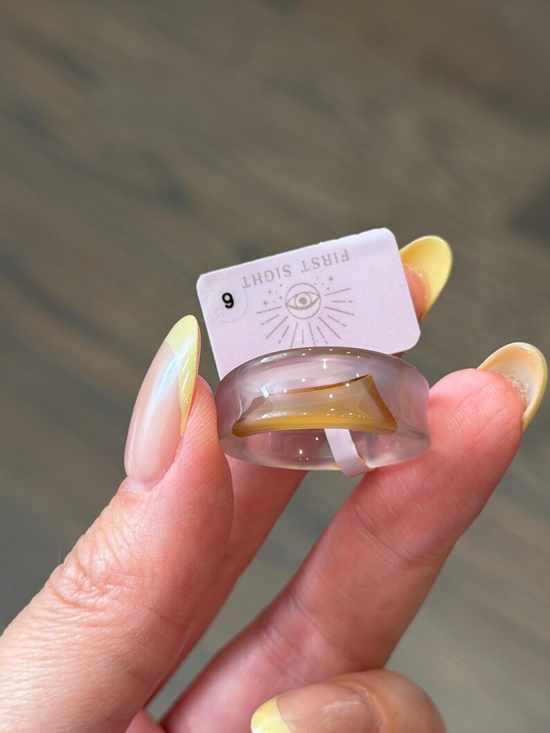 May include: A translucent light purple ring featuring an embedded amber-colored curved accent. Attached to the ring is a small rectangular tag with the text FIRST SIGHT, an eye icon, and the number 6 printed in black ink on a light pink background.