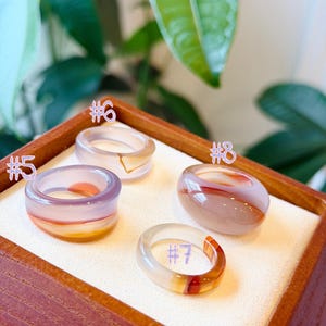 May include: Four smooth, polished stone rings labeled #5, #6, #7, and #8. Ring #5 is purple and orange, #6 is translucent with a gold accent, #7 is clear with a red band, and #8 is a rounded, opaque beige and brown stone ring.