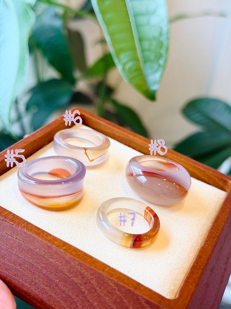 May include: Four smooth, polished stone rings labeled #5, #6, #7, and #8. Ring #5 is purple and orange, #6 is translucent with a gold accent, #7 is clear with a red band, and #8 is a rounded, opaque beige and brown stone ring.