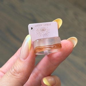 May include: A clear resin ring featuring a wavy amber-colored band detail. The ring is attached to a light pink rectangular display card printed with the text FIRST SIGHT, an eye icon with radiating lines, and the number 4 in a small circle.