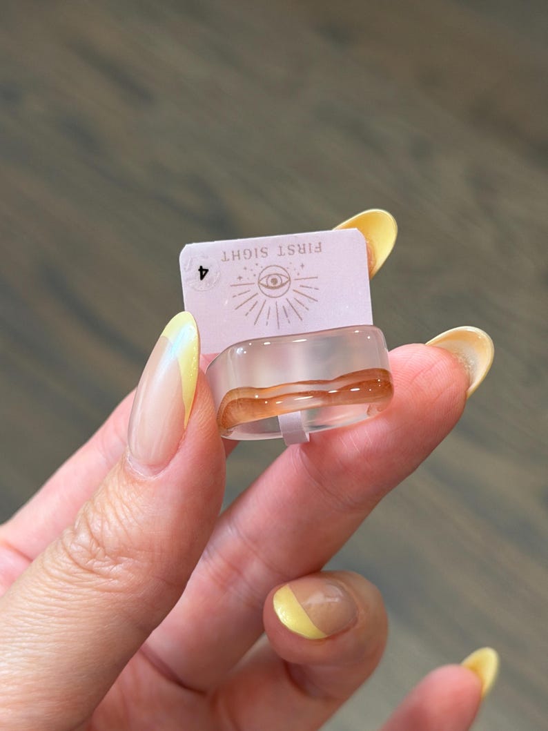 May include: A clear resin ring featuring a wavy amber-colored band detail. The ring is attached to a light pink rectangular display card printed with the text FIRST SIGHT, an eye icon with radiating lines, and the number 4 in a small circle.
