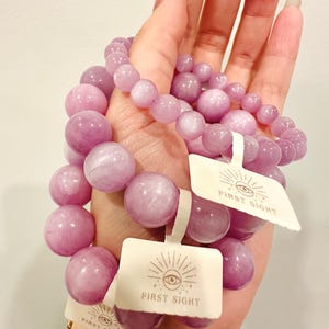 May include: Several light purple beaded bracelets with tags that read "FIRST SIGHT". The bracelets are made of round, polished beads in varying sizes. A gold chain bracelet is visible in the background.