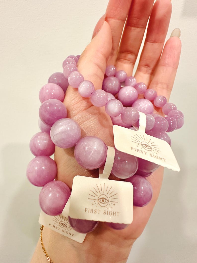 May include: Several light purple beaded bracelets with tags that read "FIRST SIGHT". The bracelets are made of round, polished beads in varying sizes. A gold chain bracelet is visible in the background.