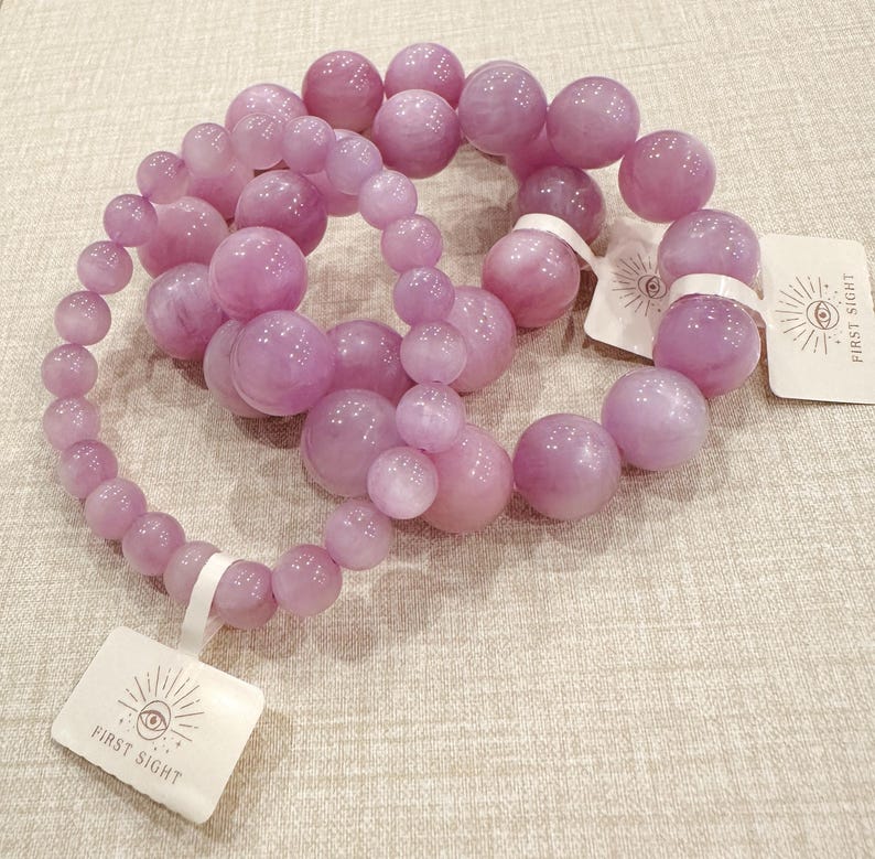 May include: A collection of light purple beaded necklaces with varying bead sizes. The beads have a glossy finish. Each necklace is attached to a white tag with the text "FIRST SIGHT" and an eye design.