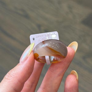 May include: A smooth, polished ring crafted from banded agate stone featuring translucent layers of gray, white, and amber-orange. A small white rectangular tag is attached to the ring, displaying the number 8 and the text FIRST SIGHT.