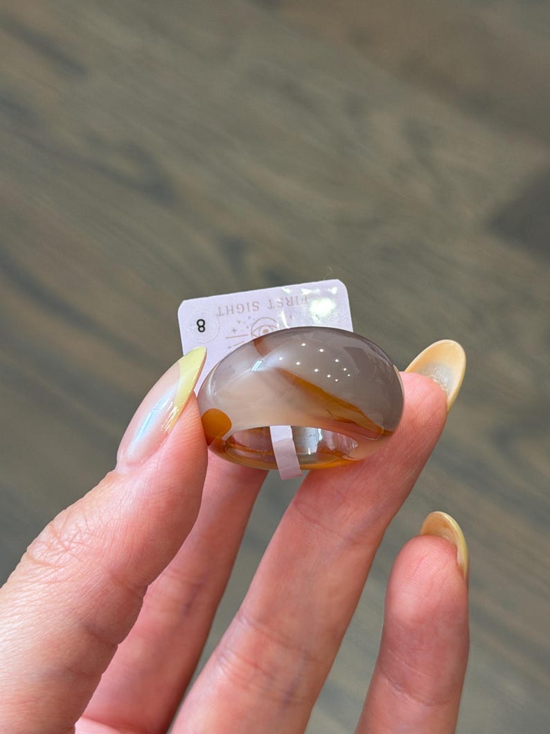 May include: A smooth, polished ring crafted from banded agate stone featuring translucent layers of gray, white, and amber-orange. A small white rectangular tag is attached to the ring, displaying the number 8 and the text FIRST SIGHT.