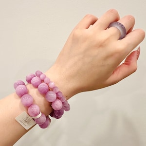 May include: Two bracelets with large, round, light purple beads and a matching ring. The bracelets are stacked on top of each other. The ring is on a finger. A tag with the text "FIRST SIGHT" is attached to one bracelet.