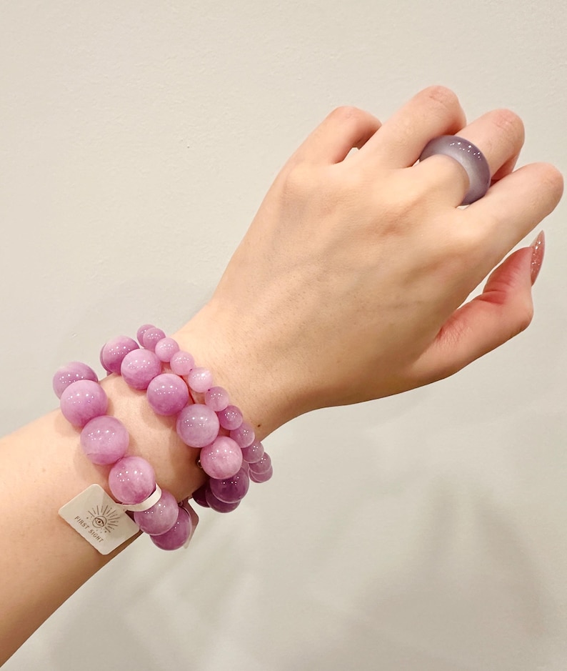May include: Two bracelets with large, round, light purple beads and a matching ring. The bracelets are stacked on top of each other. The ring is on a finger. A tag with the text "FIRST SIGHT" is attached to one bracelet.