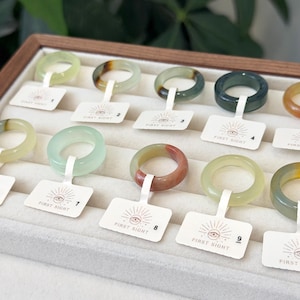 May include: Ten smooth, circular stone rings in various shades of green, yellow, and reddish-brown are displayed in a jewelry tray. Each ring features a white rectangular tag with the text FIRST SIGHT and a unique number from one to ten.