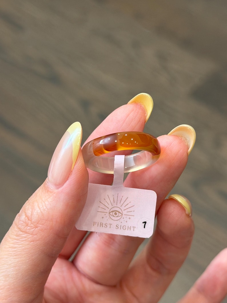 May include: A two-toned resin ring featuring a translucent amber-colored top half and a clear, colorless bottom half. A small white rectangular tag is attached to the ring, displaying the text FIRST SIGHT with an eye icon and the number 1.