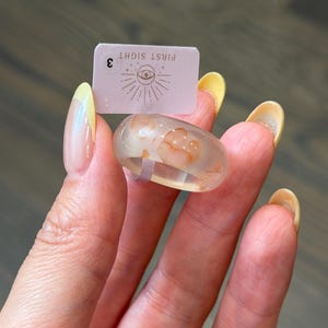 May include: A clear resin ring featuring embedded orange-toned floral or organic patterns. A small rectangular tag is positioned behind the ring, displaying the text "FIRST SIGHT" above an eye icon and the number "3" on the left side.