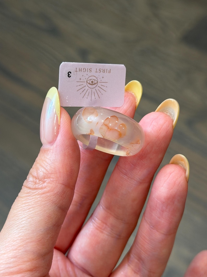 May include: A clear resin ring featuring embedded orange-toned floral or organic patterns. A small rectangular tag is positioned behind the ring, displaying the text "FIRST SIGHT" above an eye icon and the number "3" on the left side.