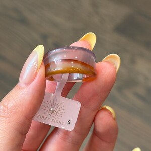 May include: A smooth, translucent band ring featuring horizontal layers of light purple and amber-brown tones. A small, rectangular white plastic tag is attached to the ring, displaying the text FIRST SIGHT and the number 5 with an eye icon.
