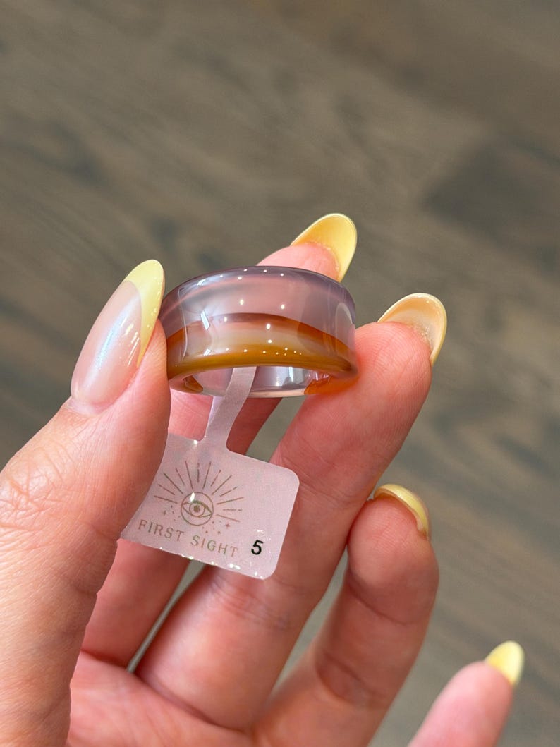 May include: A smooth, translucent band ring featuring horizontal layers of light purple and amber-brown tones. A small, rectangular white plastic tag is attached to the ring, displaying the text FIRST SIGHT and the number 5 with an eye icon.