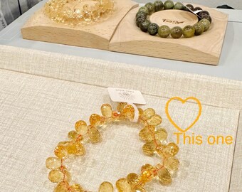 May include: A close-up of a yellow citrine gemstone bracelet with a heart drawn on the surface below. The bracelet is made of faceted, teardrop-shaped beads. The text "This one" is written on the surface below the bracelet.
