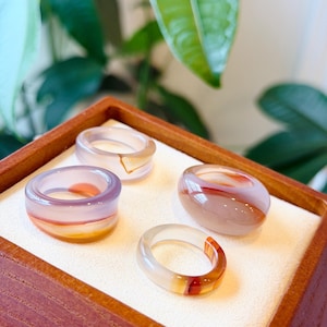 May include: Four gemstone rings in a wooden display box. The rings feature translucent bands of color, including orange, red, and purple. The rings are displayed on a white surface, with a blurred green background.