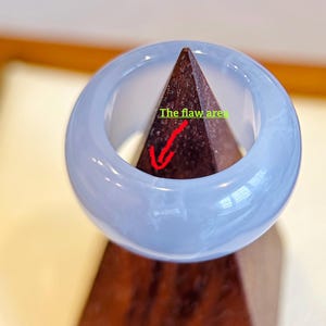 May include: A light blue jadeite bangle bracelet. The smooth, polished surface of the bracelet is a soft, translucent blue. The text "The flaw area" is visible on the image. The bangle is displayed on a brown wooden stand.