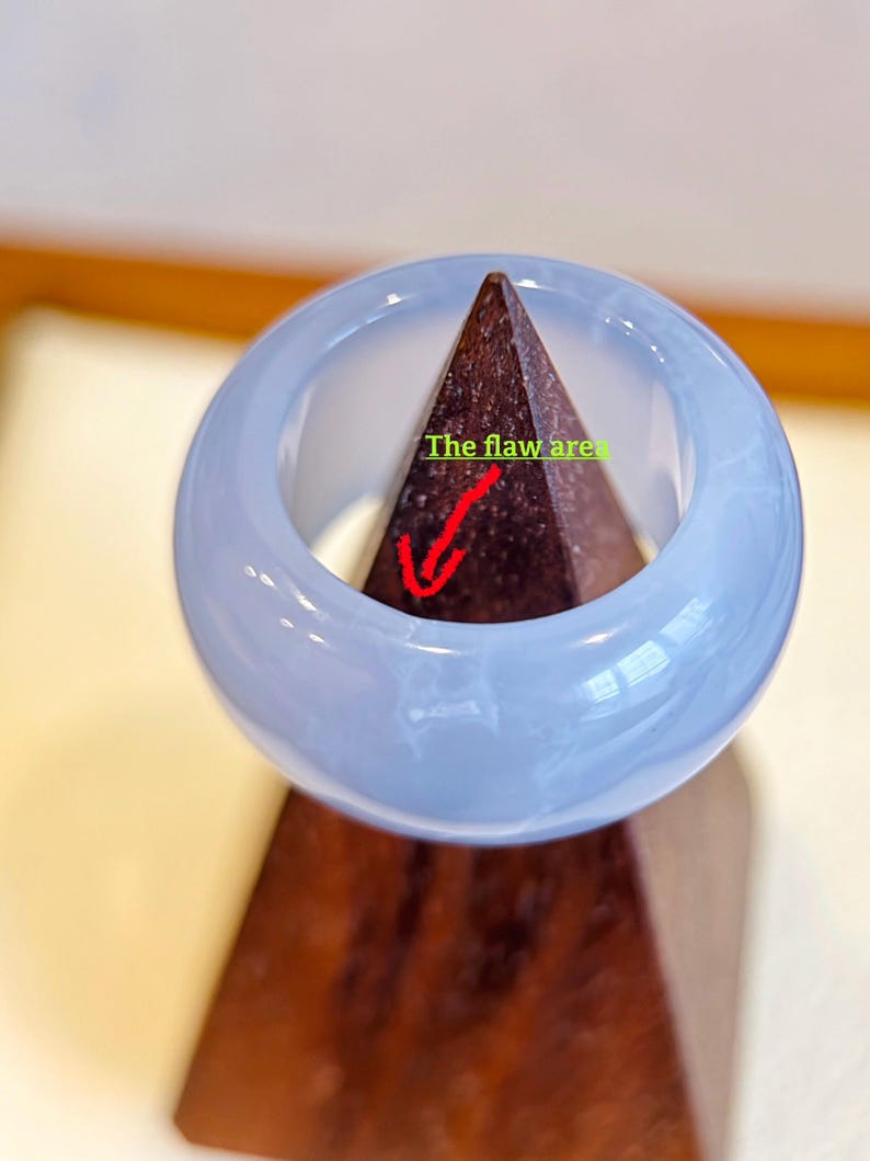 May include: A light blue jadeite bangle bracelet. The smooth, polished surface of the bracelet is a soft, translucent blue. The text "The flaw area" is visible on the image. The bangle is displayed on a brown wooden stand.