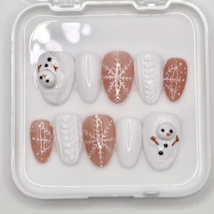 Puede incluir: Un conjunto de diez uñas postizas blancas y rosadas con diseños de temática invernal. Los diseños incluyen copos de nieve, un muñeco de nieve y un patrón de punto.