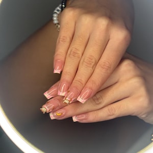 Könnte beinhalten: Manikürte Nägel mit French-Tip-Design, mit rosa und weißem Rand. Einige Nägel haben goldfarbene Affen- und Bananenanhänger. Die Nägel sind lang und oval geformt.