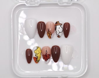 May include: Set of ten false nails with autumn-themed designs. The nails are in a clear plastic case. The designs include brown, white, and yellow leaves, a cartoon dog wearing a hat, and a white nail with no design.