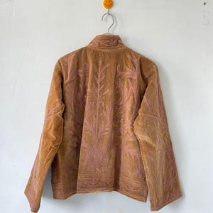May include: A tan velvet jacket featuring intricate pink floral and leaf embroidery across the back and sleeves. The jacket has a high collar, long sleeves, and a straight hemline, showcasing detailed stitching patterns throughout the fabric.