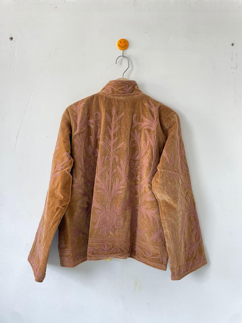 May include: A tan velvet jacket featuring intricate pink floral and leaf embroidery across the back and sleeves. The jacket has a high collar, long sleeves, and a straight hemline, showcasing detailed stitching patterns throughout the fabric.